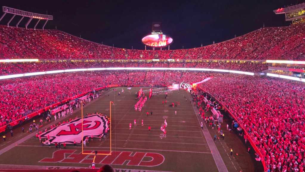 A photo of the inside of Arrowhead Stadium. A flag the shape of the Chiefs logo is on the stadium and all the lights are red.