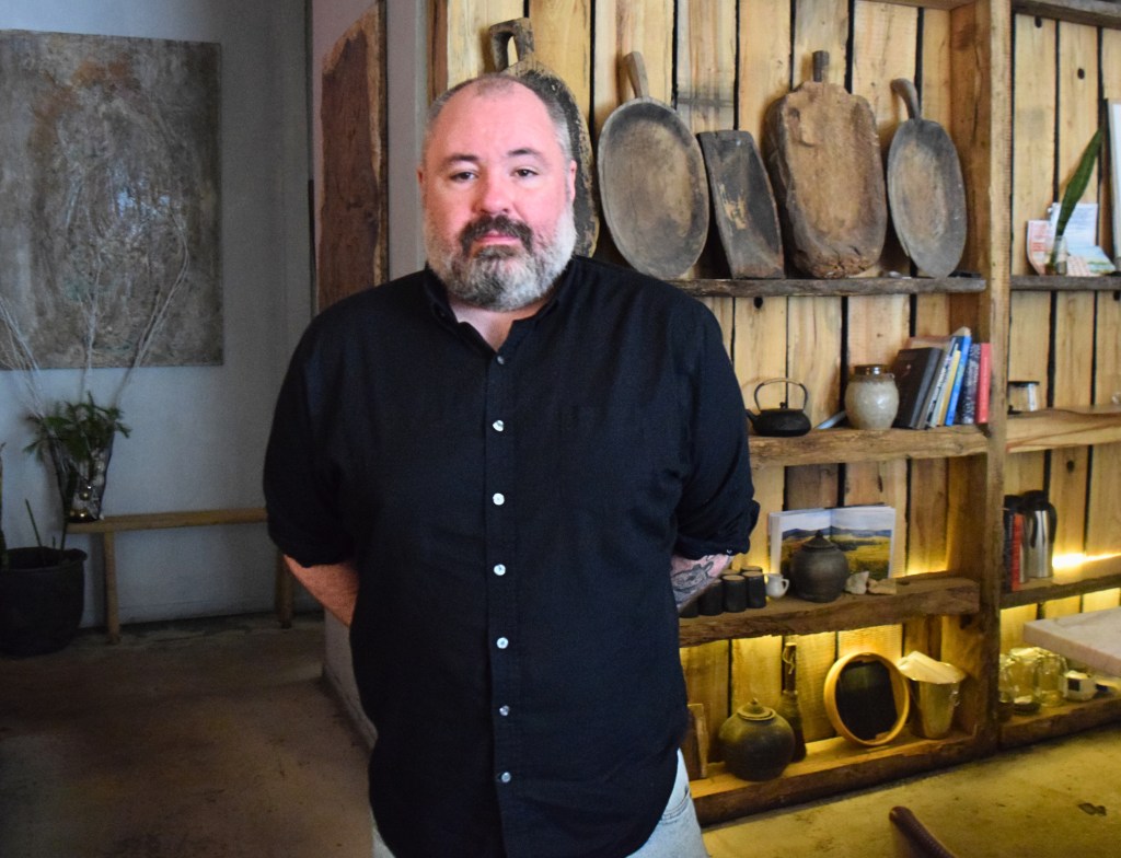 Jacob Brewer, General Manager of Noka restaurant, stands in the dining room wearing a black button-down shirt, with rustic wooden shelving displaying vintage cutting boards and pottery behind him