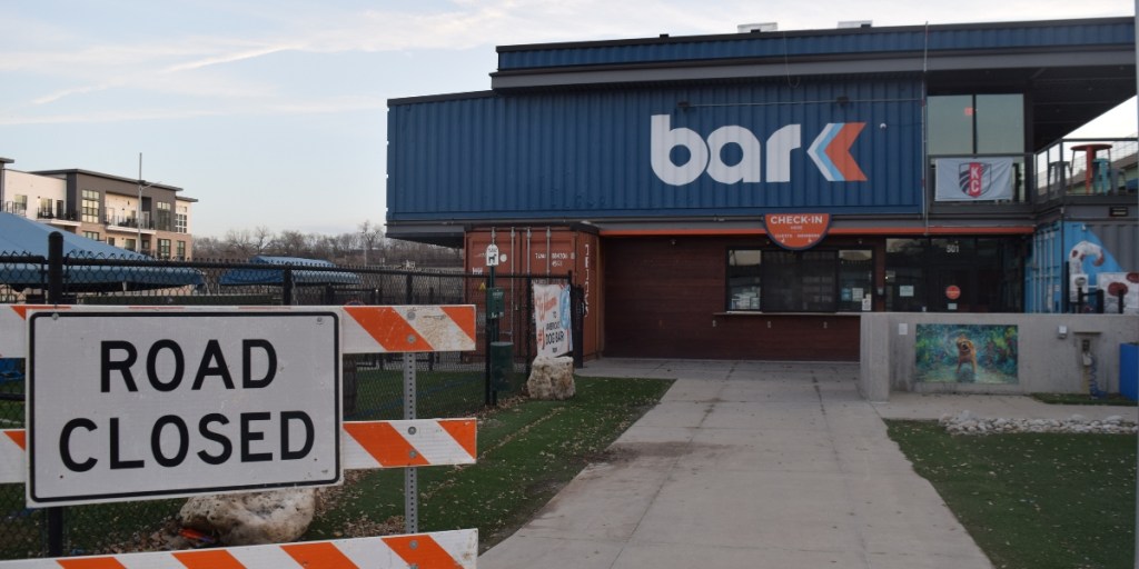 Road Closed sign in foreground with orange and white construction barriers blocking access to Bar K, a dog park and bar with blue corrugated metal exterior, in Kansas City.
