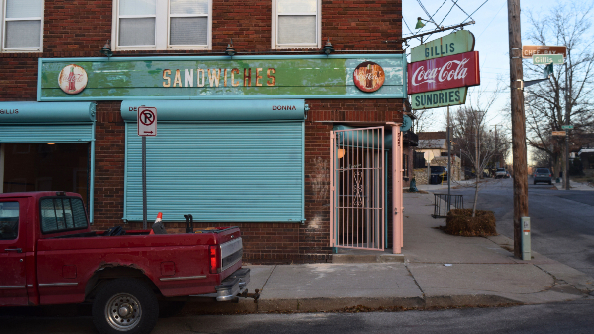 Closed storefront of Happy Gillis cafe with turquoise roll-down gate, vintage 'Sandwiches' signage and Coca-Cola Sundries sign on red brick building, with a red pickup truck parked in front