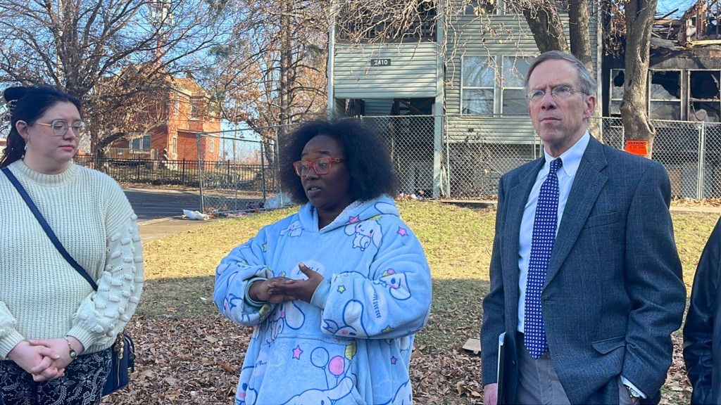Three people at outdoor press conference - woman in white sweater on left, Black woman in blue patterned hoodie speaking in center, man in business suit on right - with damaged apartment building in background