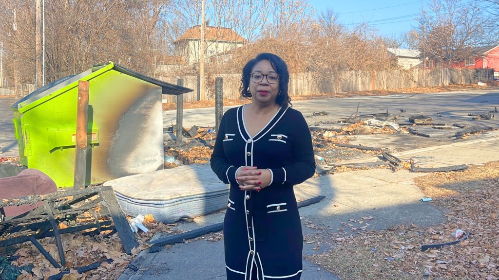 Black woman in black and white cardigan standing on sidewalk with damaged yellow-green portable structure and debris-covered lot behind her