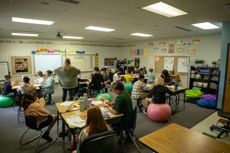 A classroom full of students work at their desks. Two teachers are helping with questions.