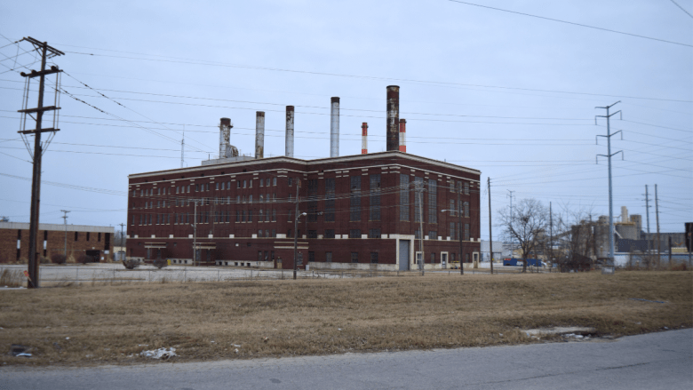 A large abandoned brick industrial building with multiple tall smokestacks against a gray sky, surrounded by vacant land and utility poles.