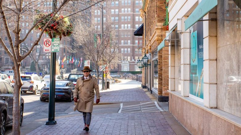 A man wearing a coat walking down the sidewalk in the Plaza, with South Plaza apartments and Brush Creek in the background