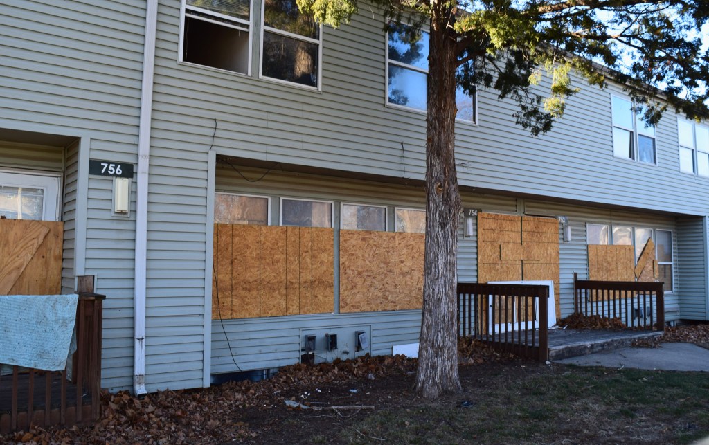 Two-story light-colored apartment building with plywood boards covering first-floor windows, unit numbers 756 and 754 visible, residential neighborhood setting