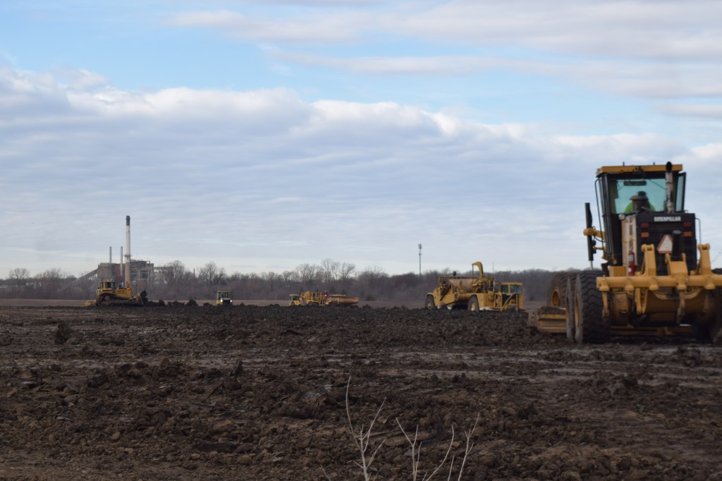 A row of large yellow Caterpillar graders and scrapers moves earth across a wide, muddy field on an overcast day. The smokestack and structure of the Blue Valley Power Plant are visible on the horizon to the left. Bare trees line the distant treeline. A Caterpillar motor grader is prominently visible in the foreground on the right.