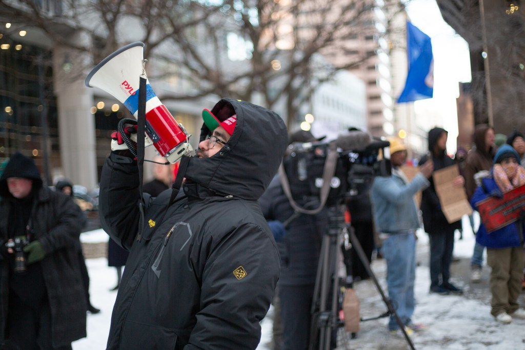 A protestor, bundled up in a winter coat, holding a megaphone, to the left of a crowd holding signs.