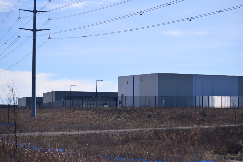 Three large, boxy data center buildings with gray paneling sit behind a security fence amid scrubby vegetation. High-voltage power transmission lines cross the foreground against a blue sky.