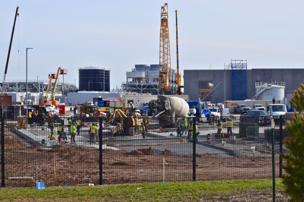 Dozens of construction workers in yellow safety vests work around heavy equipment and a concrete mixer truck at an active job site. Large cranes and a partially completed data center building are visible in the background.