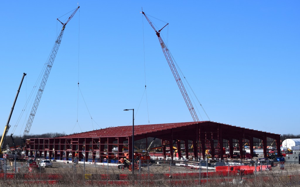 Three tall lattice cranes rise above a large partially-constructed steel-framed building against a clear blue sky. The exposed red structural steel frame spans a broad footprint, with construction equipment and vehicles visible along the perimeter.