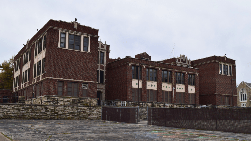 A wide-angle photograph of the vacant Bryant School, a two- and three-story red brick building with limestone accents and decorative panels along the facade. The building sits behind a stone retaining wall and chain-link fence topped with brown privacy screening. A paved lot in the foreground shows cracks and faded chalk drawings. A church steeple is partially visible on the right side of the frame. The sky is overcast.