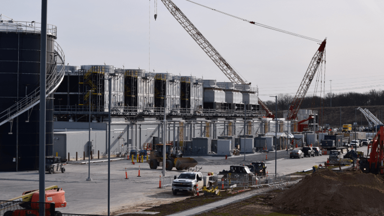 A large active data center construction site with a red crawler crane dominating the skyline. Multi-level steel and mechanical structures are visible alongside construction vehicles and workers on a paved lot.