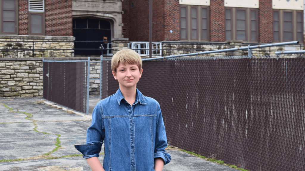 A young woman with short blond hair wearing a denim button-up shirt stands on a cracked asphalt lot in front of the vacant Bryant School. Behind her, the school's stone and brick facade is partially visible through a brown privacy fence attached to a chain-link barrier. Green moss grows in cracks in the pavement.
