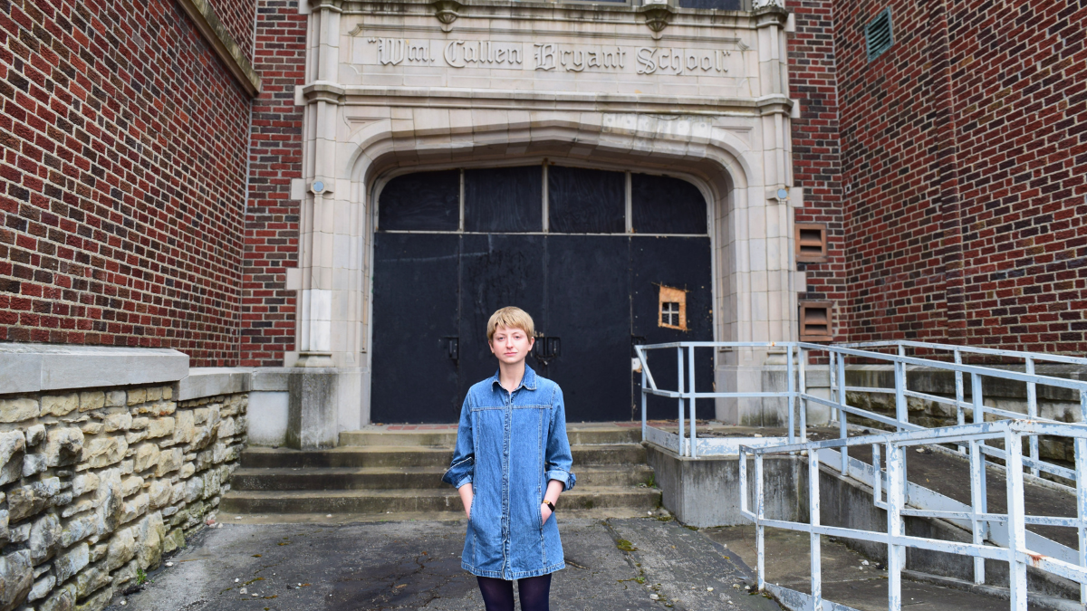A young woman with short blond hair wearing a denim shirt dress and dark tights stands at the bottom of concrete steps leading to the main entrance of the William Cullen Bryant School. The entrance archway above her reads "Wm. Cullen Bryant School" in carved stone lettering. The doors behind her are covered with dark plywood boards, one of which has a small broken-out section. A metal handicap ramp railing is visible to the right.