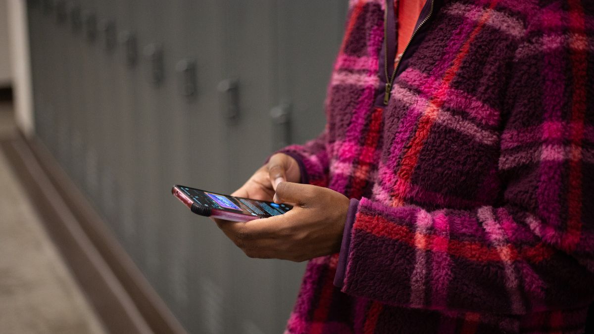 A student stands in front of lockers in a school hallway. They are looking down at their phone.