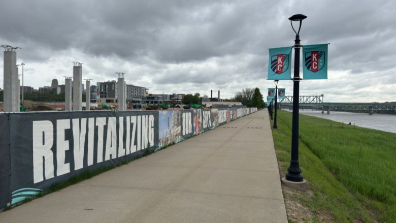 A sidewalk running alongside the Missouri River at the Berkley Riverfront. To the left is a banner that says "Revitalizing our city's riverfront," and to the right is a streetlight with the KC Current logo.
