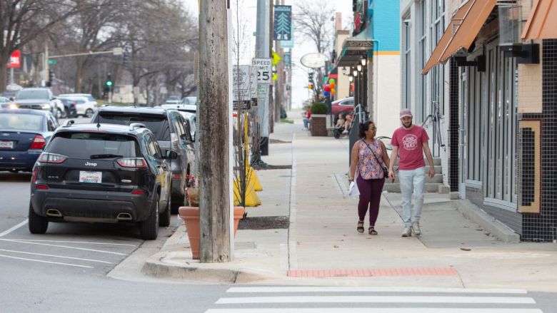 Two people walk down the sidewalk, with a line of parked cars on the street to their right and a row of storefronts on their left.