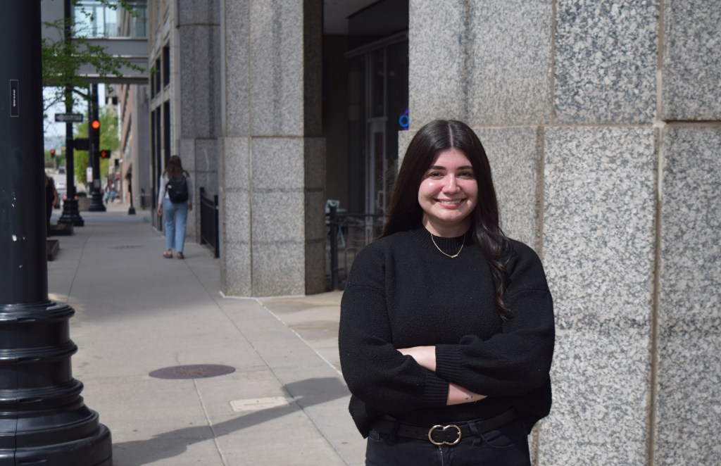 Emma Shoemaker, a young woman with long dark hair wearing a black sweater and gold necklace, stands with her arms crossed and smiling on a downtown Kansas City sidewalk in front of a granite-faced building.