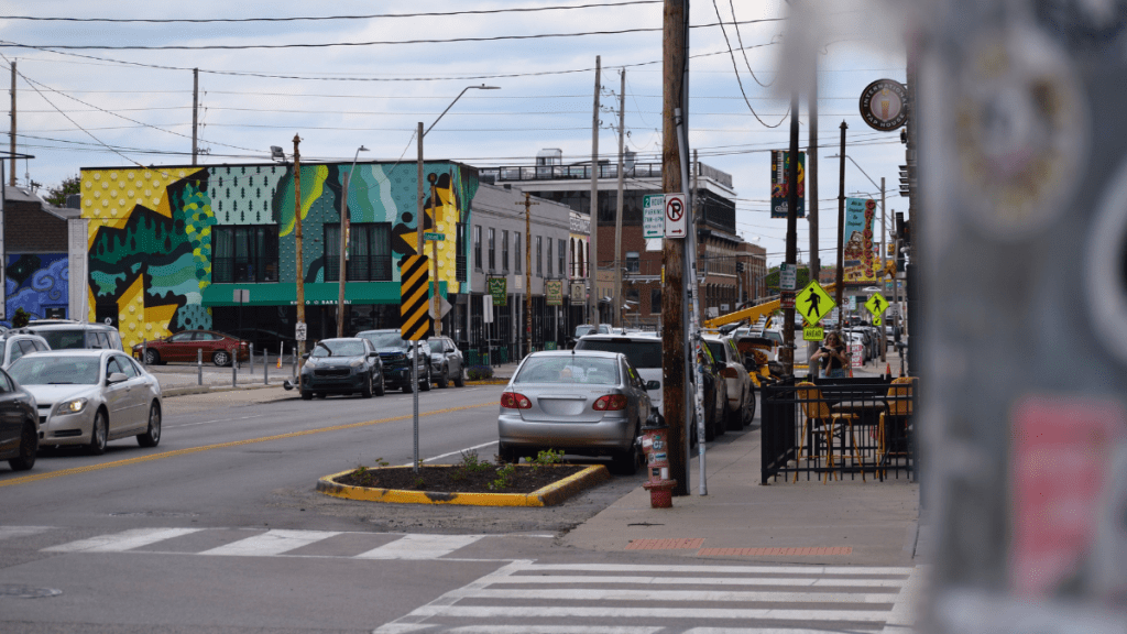 A street-level view of the Crossroads Arts District in Kansas City showing a colorful geometric mural on a building facade, parked cars along the street, outdoor restaurant patio seating, and various business signs stretching down the block under an overcast sky.
