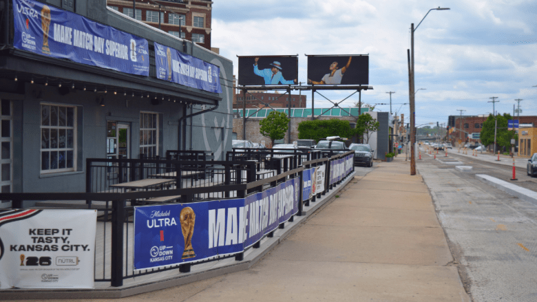 A gray-painted bar exterior with an extended outdoor patio lined with blue FIFA World Cup 2026 and Michelob Ultra promotional banners. Billboards and orange construction cones are visible along the street under a cloudy sky.