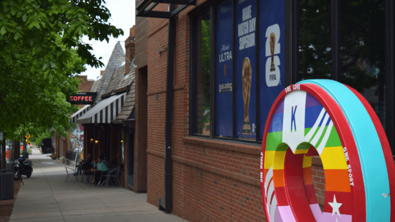 A colorful rainbow "KC heart" sculpture commemorating the FIFA World Cup 2026 sits on the sidewalk outside a brick building with World Cup promotional posters in the windows. A coffee shop with outdoor seating is visible down the block in the Westport entertainment district.