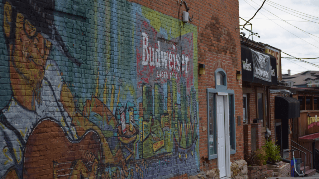 A large colorful mural featuring a guitar player and a Budweiser logo covers the brick exterior wall of a building in the Westport entertainment district. A bar entrance and signage are visible to the right.