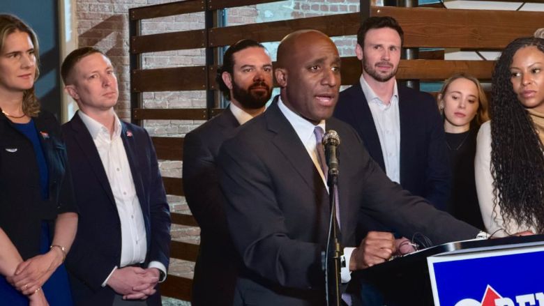 Mayor Quinton Lucas stands at a podium with a blue election sign on it. In the background are six other people, among them City Councilmembers Lindsay French and Johnathan Duncan, Lucas's chief of staff Reid Day and Jackson County Prosecutor Melesa Johnson.