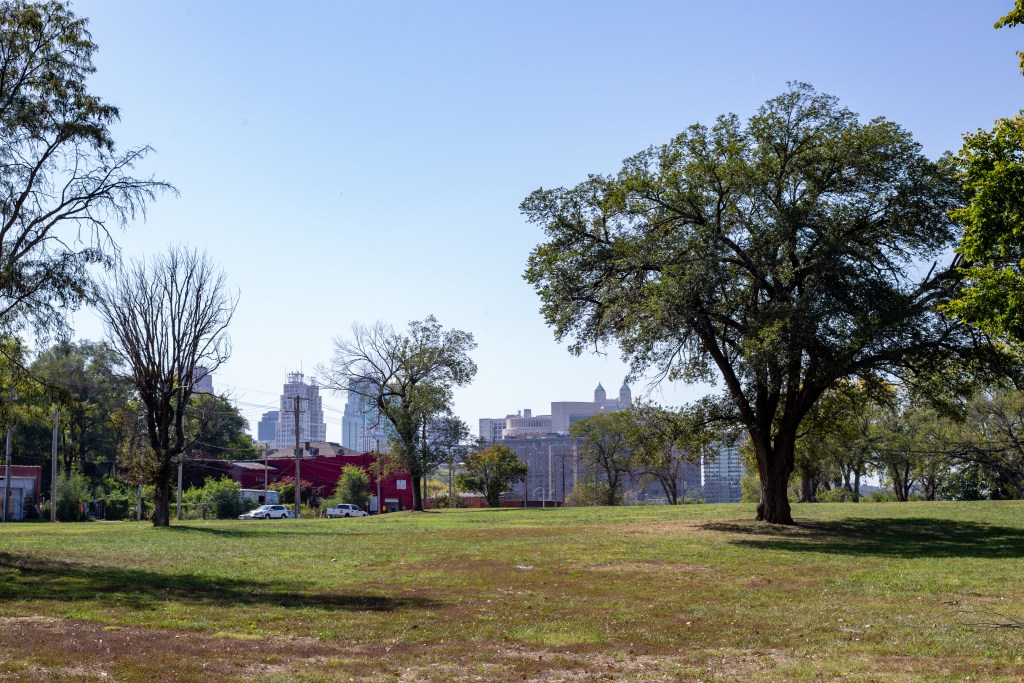 A large open park space with a mix of green and brown grass. Trees frame the photo with the Kansas City, Missouri, downtown skyline in the background before giving way to a blue sky.