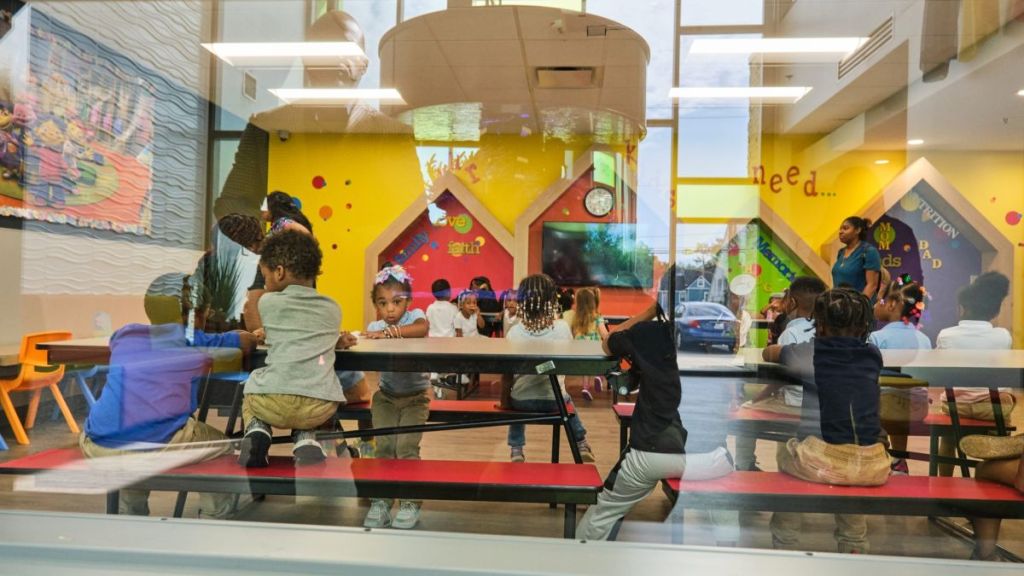 Schoolkids sitting at tables seen through a window with a reflection of a man.