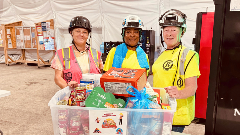 Three women in hard hats and high-visibility safety vests smile behind a large clear bin filled with donated food items including canned goods, pasta, and snacks.