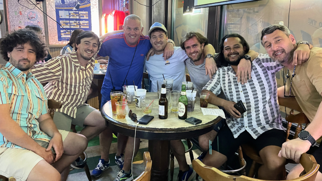 Seven men in casual summer shirts and shorts crowd shoulder to shoulder around a small round bar table, arms over one another's shoulders and grinning at the camera, with pint glasses, plastic Quaff cups, a Yuengling Lager bottle, sunglasses and phones spread across the tabletop in a dim, memorabilia-covered bar.