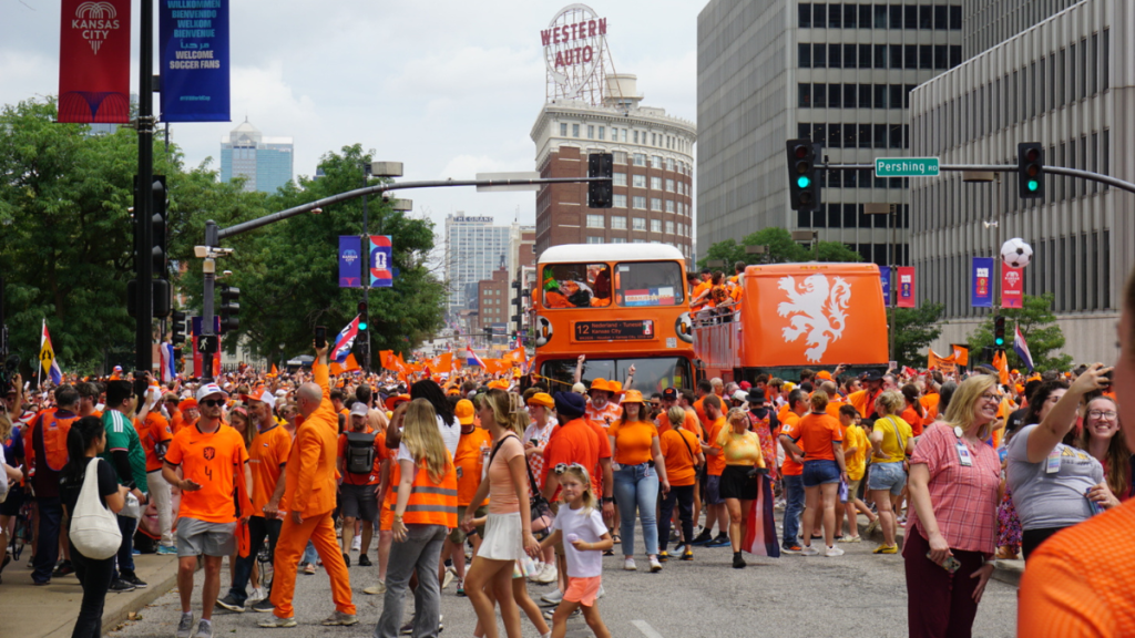 A dense crowd of soccer fans dressed almost entirely in orange fills a downtown Kansas City street, waving orange flags and following an orange double-decker bus and an orange truck bearing the Dutch lion emblem; the "Western Auto" rooftop sign and "Welcome Soccer Fans" banners rise in the background under an overcast sky.