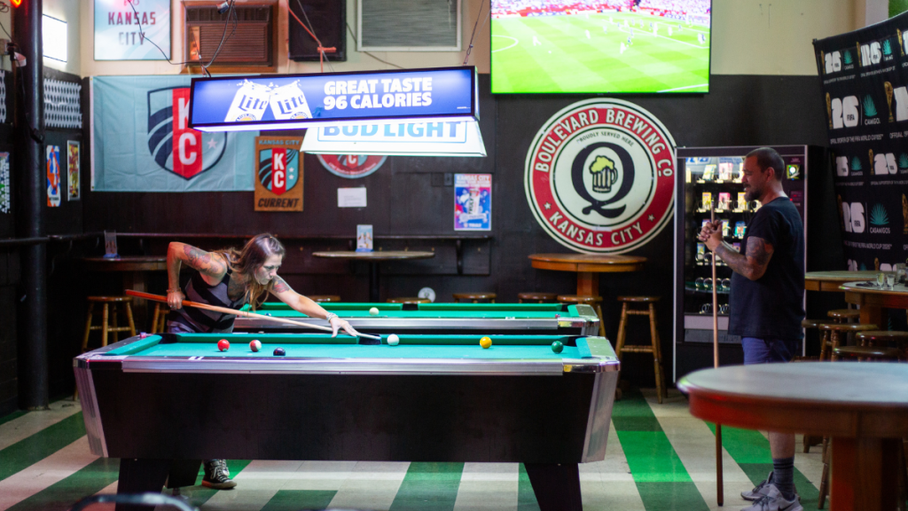 A tattooed woman leans low over a green-felt pool table to line up a shot with her cue while a man stands holding his cue at the far end; above them a lit Miller Lite and Bud Light fixture and a soccer match on a wall-mounted TV glow over a round Boulevard Brewing Co. "Q" sign, a vending machine and a green-and-white checkered floor.