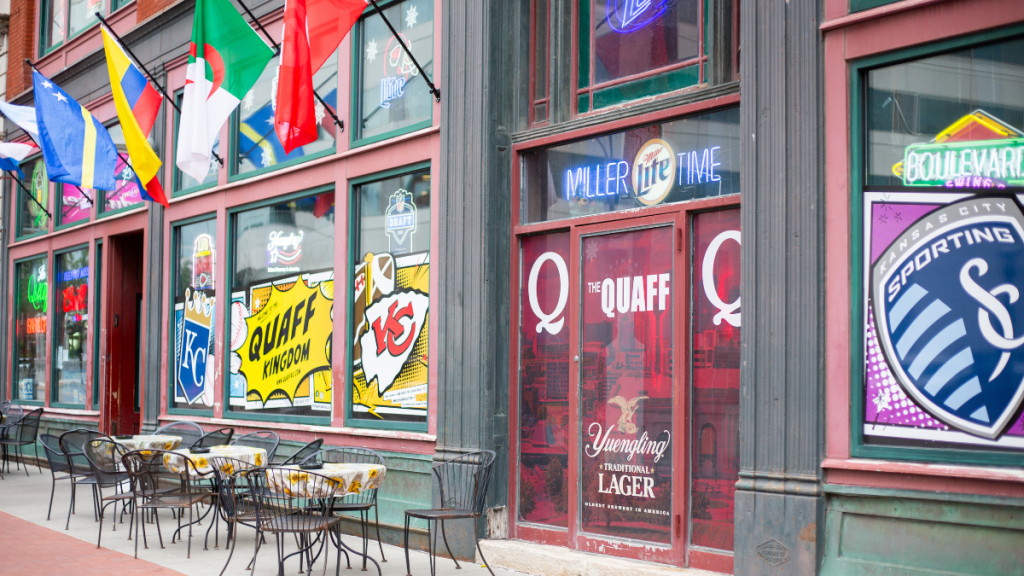 A close view of The Quaff's street corner, with "The Quaff" lettering on a red entry door beneath a "Miller Lite Time" neon sign, ground-floor windows painted with "Quaff Kingdom," a Royals logo, a Sporting KC crest and a Yuengling Lager ad, national flags projecting overhead, and an empty sidewalk patio of metal tables and chairs with sunflower-print cloths.