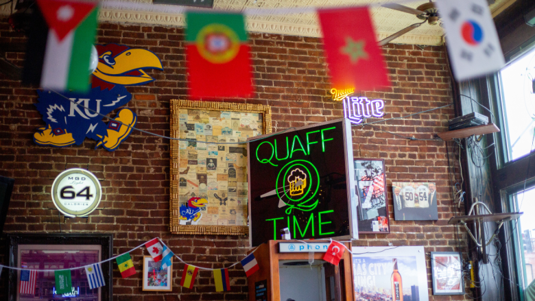 Inside a bar, strings of small national flags hang across an exposed red brick wall covered in neon signs — including a bright green "Quaff Q Time" clock sign with a beer mug, a Miller Lite sign and an MGD Light 64 sign — alongside a University of Kansas Jayhawk cutout, framed photos and a cork-bordered collage, beneath a pressed-tin ceiling.