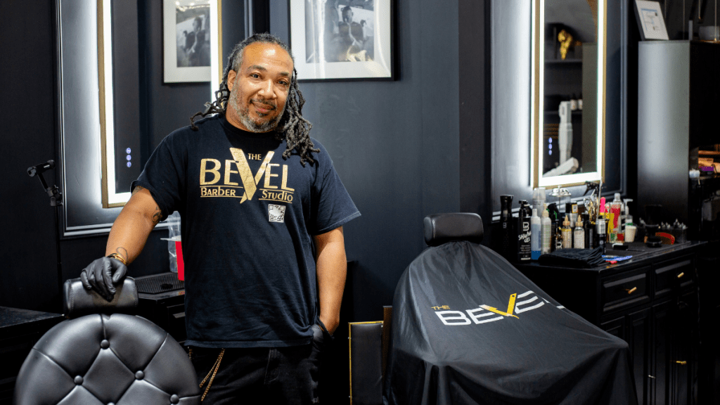 Casie Murff, a barber with long locs and a gray-flecked beard, stands smiling in a black "The BeVel Barber Studio" T-shirt and black gloves, one hand resting on a black-and-gold barber chair in his dark, mirror-lined shop.