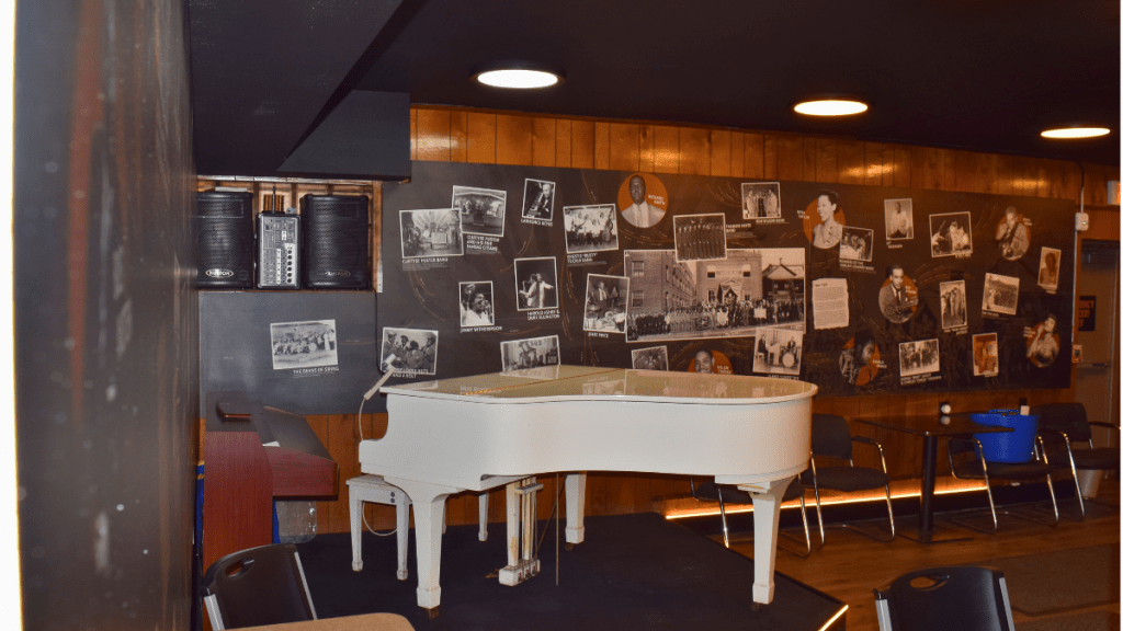 A white grand piano stands on a low stage inside the Mutual Musicians Foundation in Kansas City's 18th and Vine Jazz District. Behind it, a dark wall displays a large collage of black-and-white photographs of jazz musicians and bands connected to the foundation's history.