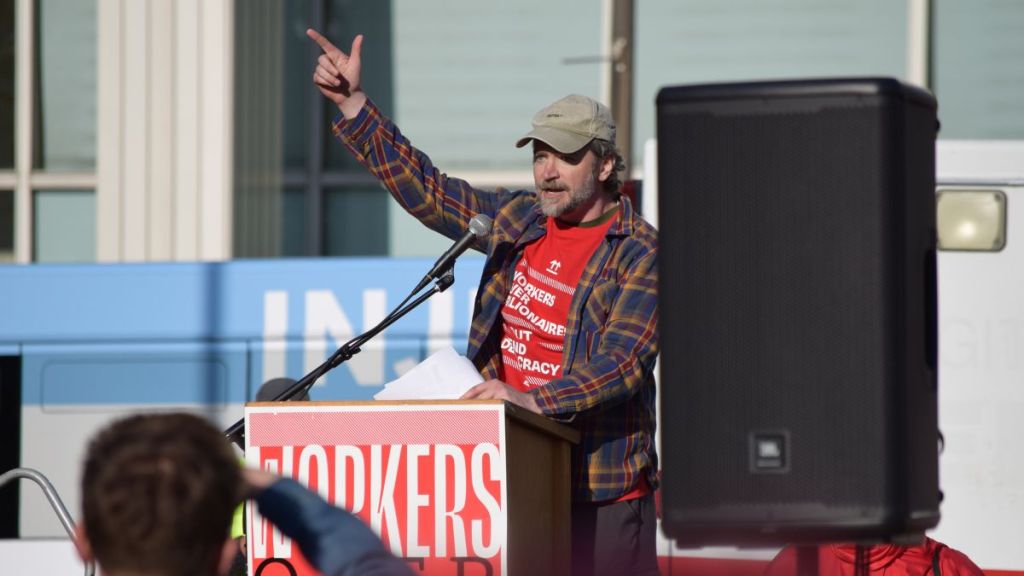 A man wearing a red Missouri Workers Center with a plaid jacket over top speaks into a microphone on a podium at a rally