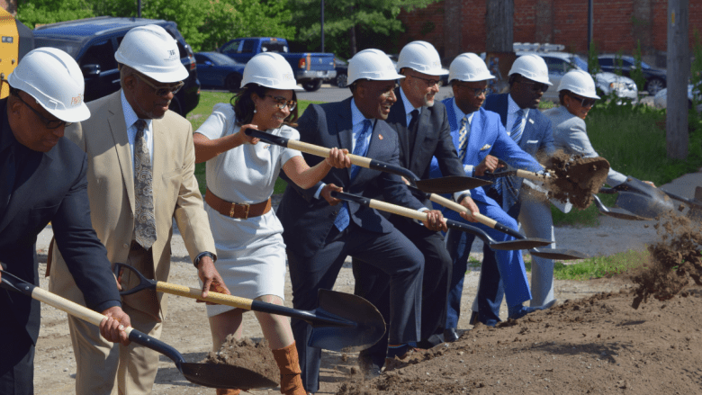 A row of nine people in white hard hats and business attire lean forward in unison, pushing shovels into a mound of dirt at a groundbreaking ceremony. Loose soil flies from the shovels of those at the right end of the line. Parked cars, trees and a brick building are visible in the background.
