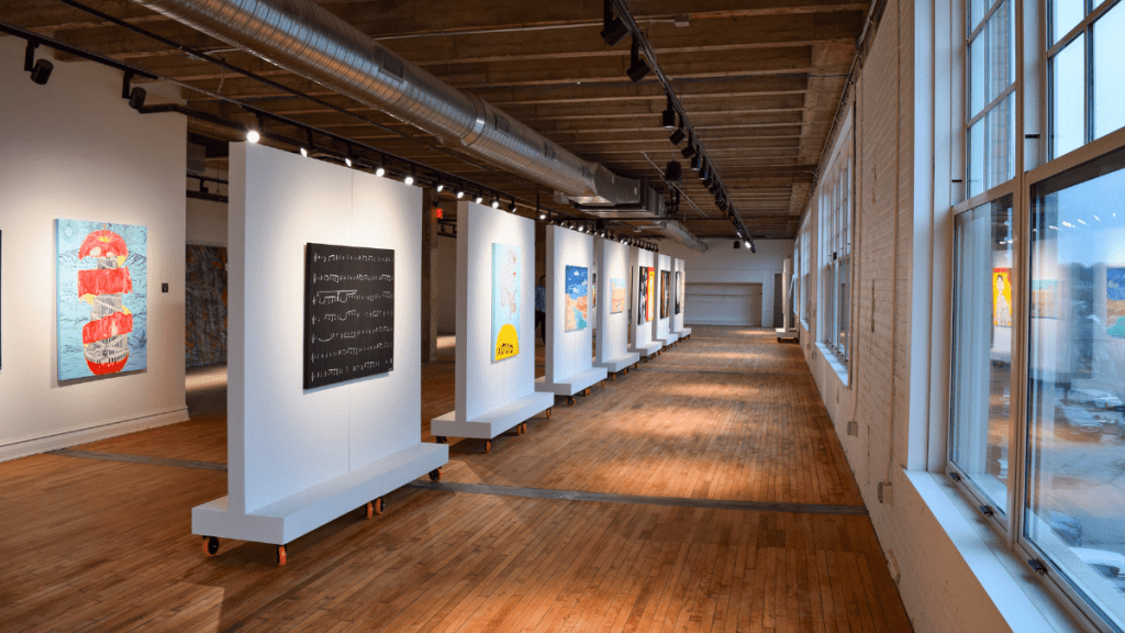 A long, sunlit gallery with wide-plank wood floors and an exposed timber ceiling. Paintings hang on a row of freestanding white partition walls on rolling casters that recede down the center of the room, with more artwork on the far wall. Tall windows line the right side, and track lighting runs overhead.