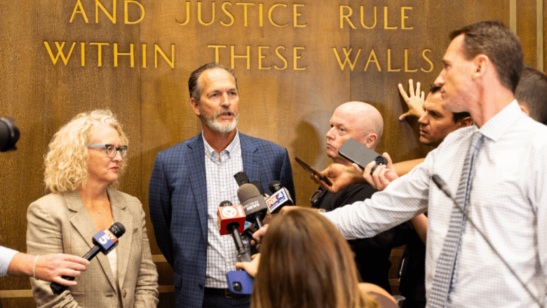 A man in a blue plaid blazer and checked shirt speaks into a cluster of television microphones while a woman in a tan plaid blazer stands beside him, listening. Reporters crowd in from the right holding phones and recorders. Behind them, gold lettering carved into a wood-paneled wall reads "AND JUSTICE RULE WITHIN THESE WALLS."