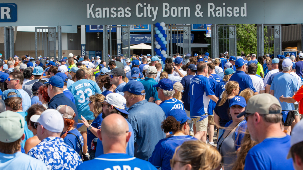 A dense crowd of fans in blue and light blue Royals jerseys, T-shirts and caps walks toward a stadium entrance on a bright day. A jersey in the foreground reads "GORDON." Large lettering above the gates reads "Kansas City Born & Raised," with a column of blue and white balloons beneath it.