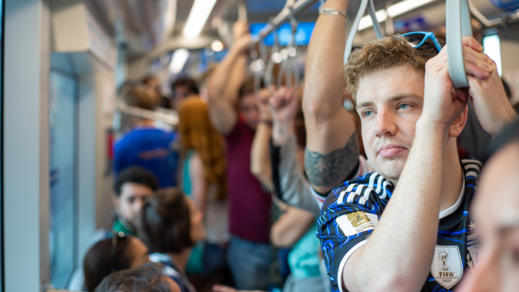 A crowded streetcar car interior with riders standing and holding overhead straps; in the foreground, a man wearing a blue and black soccer jersey with a FIFA World Cup patch looks toward the window.