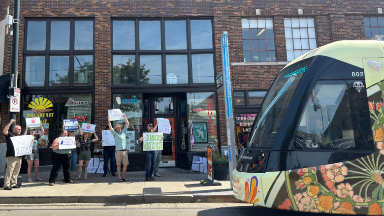 A group of people lined up stand on a sidewalk outside a brick storefront with "Breakfast," "Shakes" and "Coffee" signage, raising hand-lettered signs supporting the streetcar workers' union vote, as a floral-painted Kansas City Streetcar passes in the foreground.