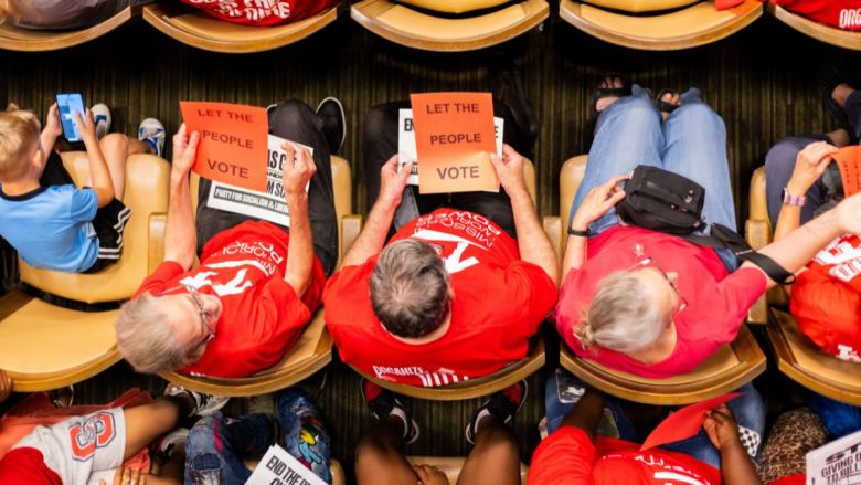 Overhead view of people wearing red Missouri Workers Power t-shirts, holding paper signs that say "Let the People Vote"
