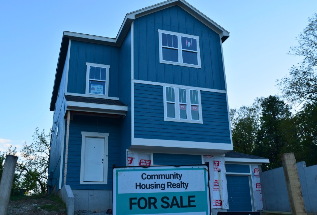 A tall, narrow blue three-story house with white trim nears completion. The upper floors have vertical board-and-batten siding and the lower floors have horizontal lap siding. A white front door sits raised above a bare concrete foundation. A "Build WyCo" sign hangs in an upper window, and part of the ground floor is still covered in house wrap beside a small attached garage with a blue door. A teal and white "Community Housing Realty For Sale" sign stands in the foreground against a dusk sky and trees.