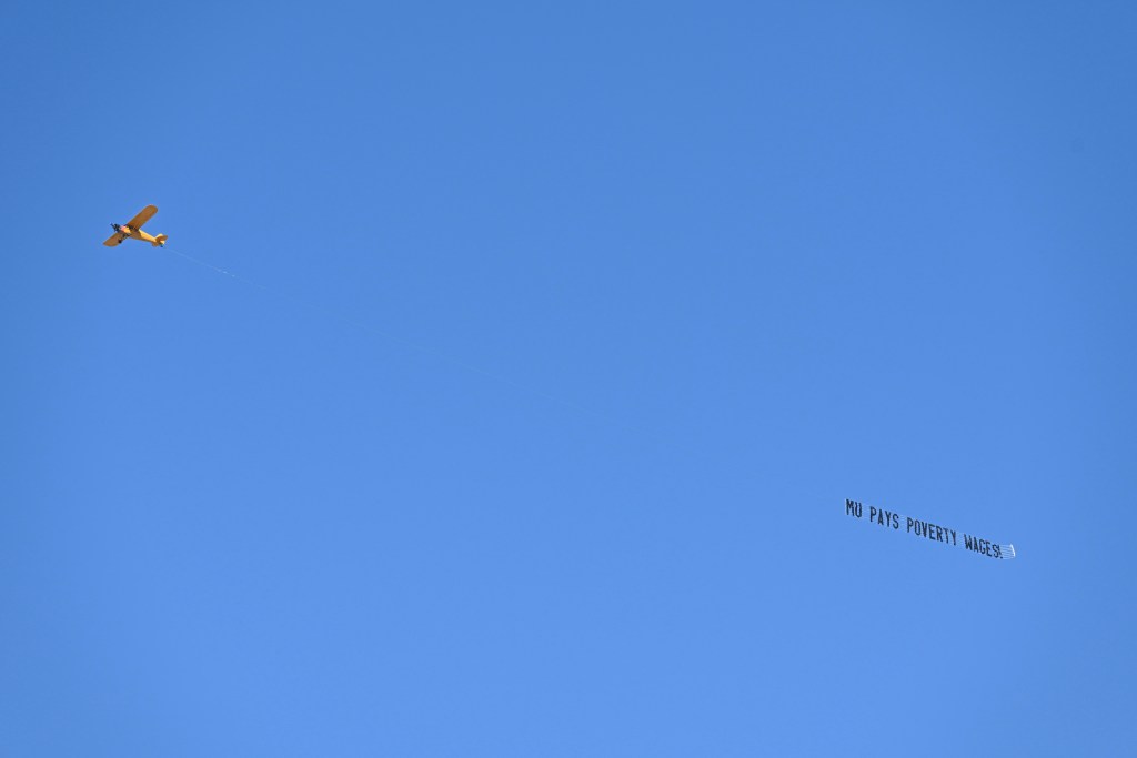 A yellow single-engine plane flies against a clear blue sky, towing a long banner reading "MU pays poverty wages!"