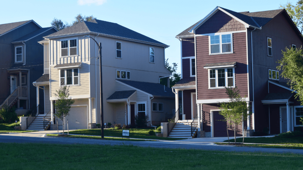 Two new three-story homes side by side on a residential street, one beige and one maroon, both with gray shingled roofs. Each has a front-facing garage at street level and a staircase to a covered front porch, plus a lower wing to the side with its own entrance. Young trees, fresh lawns and a sidewalk line the front of the homes, with a similar dark gray house partly visible at left.