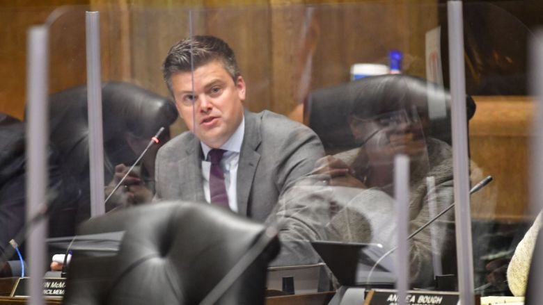 City Councilmember Kevin McManus speaks into a microphone behind a COVID-lockdown-era plexiglass shield
